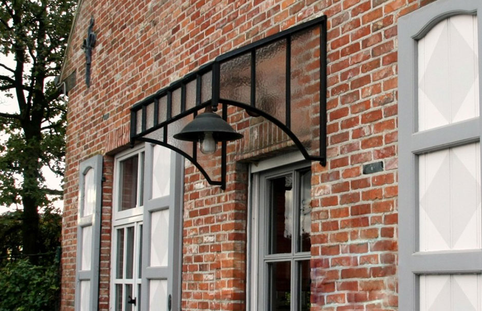 Vordach aus Glas und Metall für Haustüren – modernes Design, wetterfest und stabil Vordach aus Glas und schwarzem Metall über einer Haustür an einer Backsteinfassade mit weiß-grauen Fensterläden