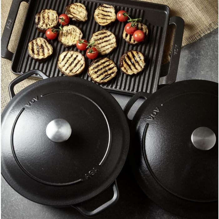 Gusseisen-Kochgeschirr-Set mit Bratplatte, Grillgemüse und Tomaten – Hochwertige Töpfe für gesundes Kochen Zwei gusseiserne Kochtöpfe mit Deckel und eine Grillplatte mit gegrillten Auberginenscheiben und Kirschtomaten auf einem Küchentisch