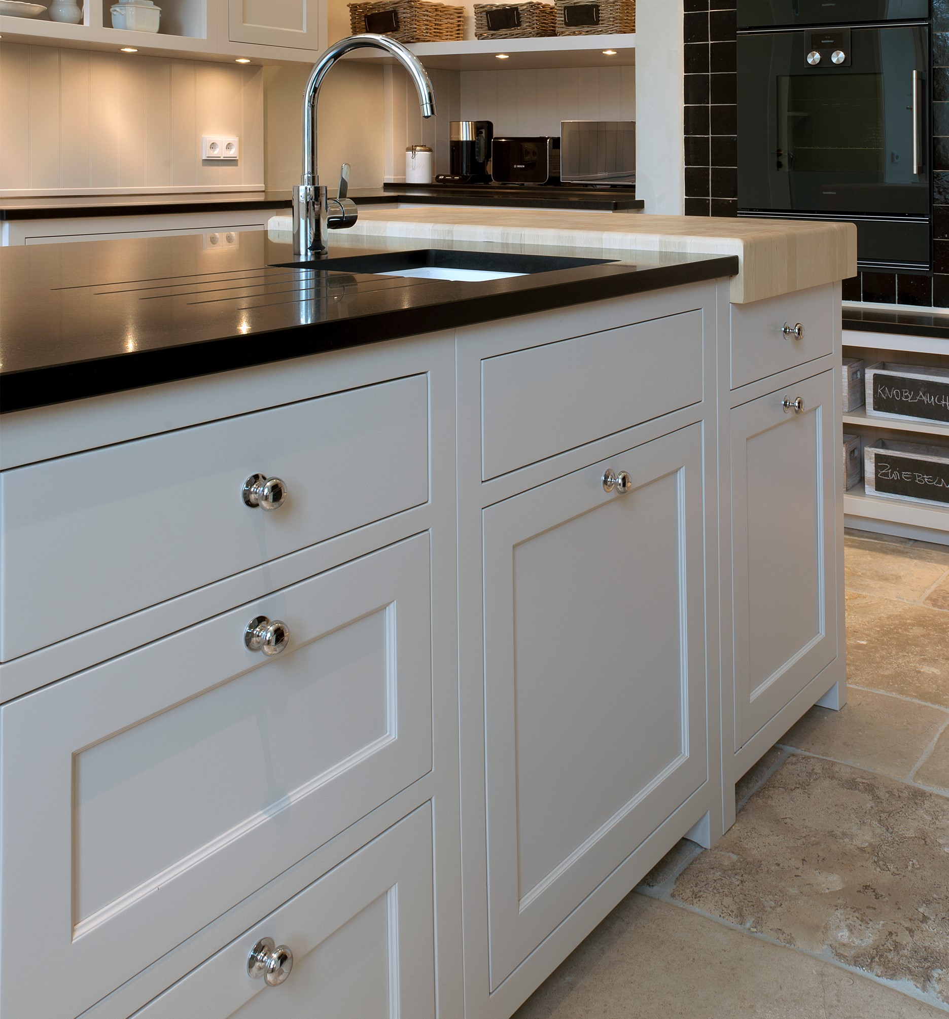 Helle Kücheninsel mit Shaker-Fronten, schwarzer Stein-Arbeitsplatte, Unterbauspüle, Chrom-Armatur und Holz-Arbeitsblock; Blick auf Schubladen mit Knöpfen und Natursteinboden