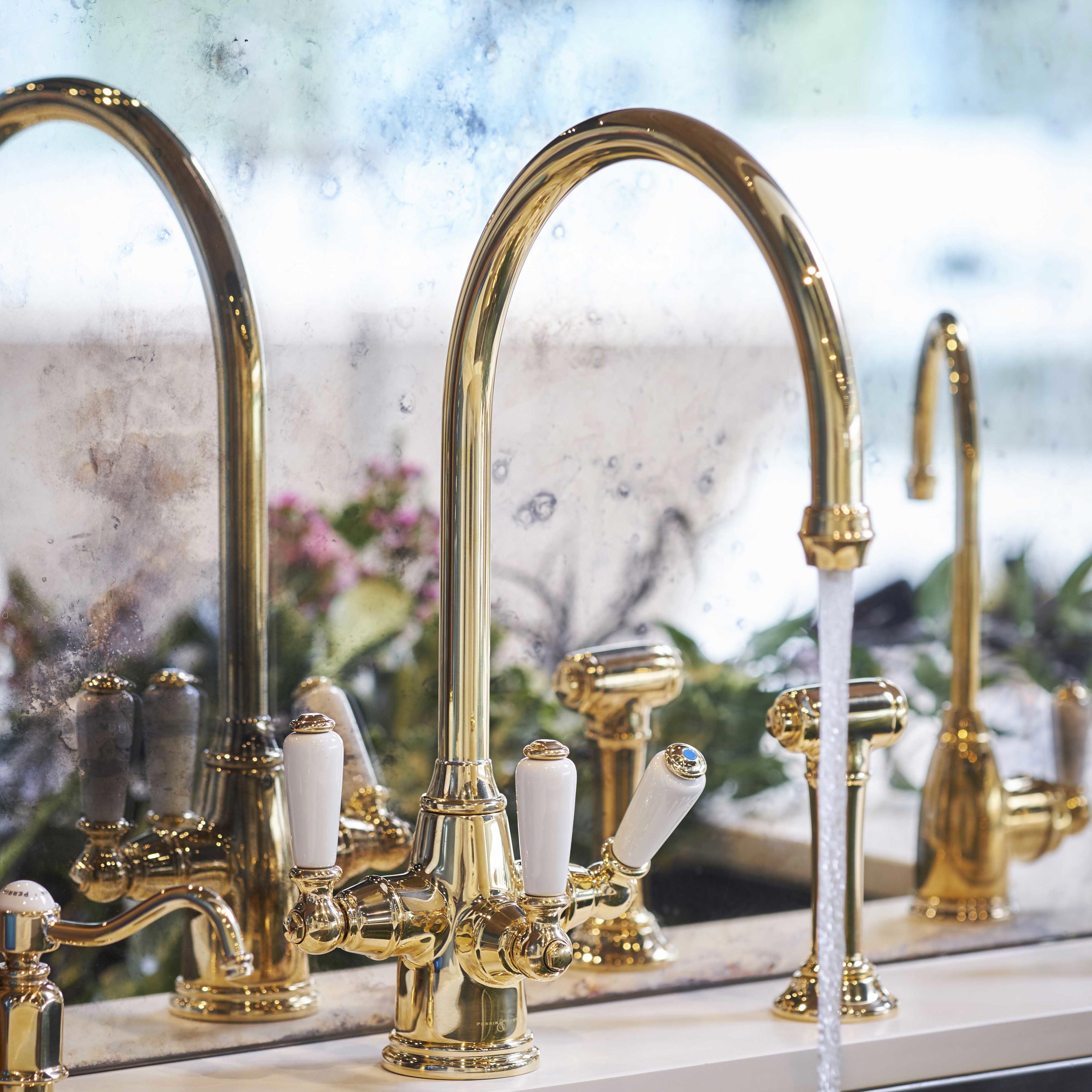 Elegante goldene Küchenarmatur mit hohem Auslauf, weißen Keramikhebeln und seitlicher Handbrause; Wasser läuft, Spiegel im Hintergrund