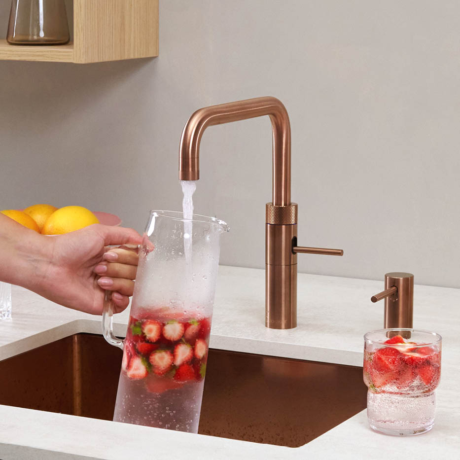 Moderne Küchenarmatur in Kupferoptik beim Befüllen einer Karaffe mit Wasser und Erdbeeren Hand hält eine Glaskaraffe mit Erdbeeren unter einen Kupfer-Wasserhahn in einer modernen Küche