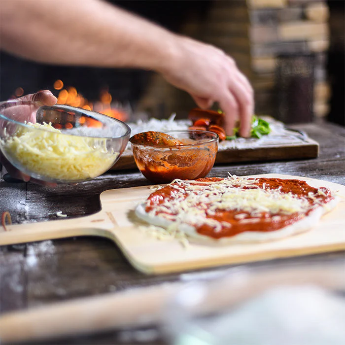 Frische Pizza wird mit Tomatensauce und geriebenem Käse vor dem Backen belegt Hand belegt frischen Pizzateig mit Tomatensauce und geriebenem Käse auf einem Holzbrett, Zutaten im Hintergrund