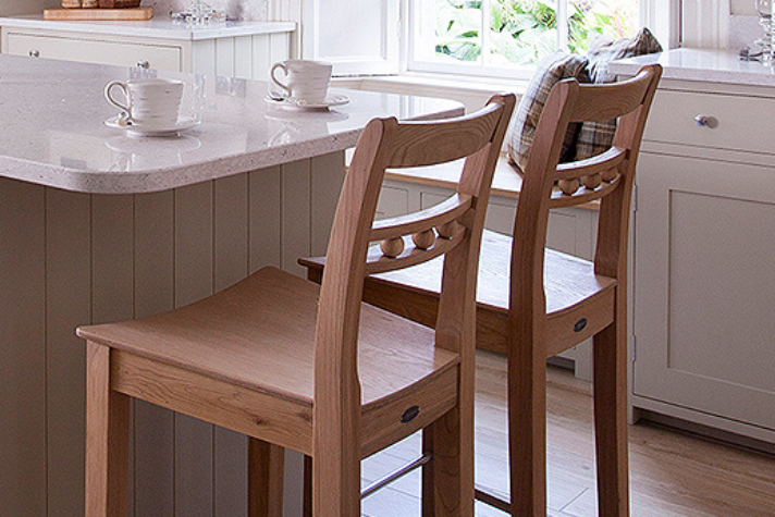 Zwei elegante Barhocker aus Holz an einer modernen Kücheninsel mit zwei Tassen auf der Arbeitsplatte Zwei Holz-Barhocker stehen an einer hellen Kücheninsel, auf der zwei Tassen und Untertassen stehen, im Hintergrund sind ein Fenster und Küchenschränke sichtbar.