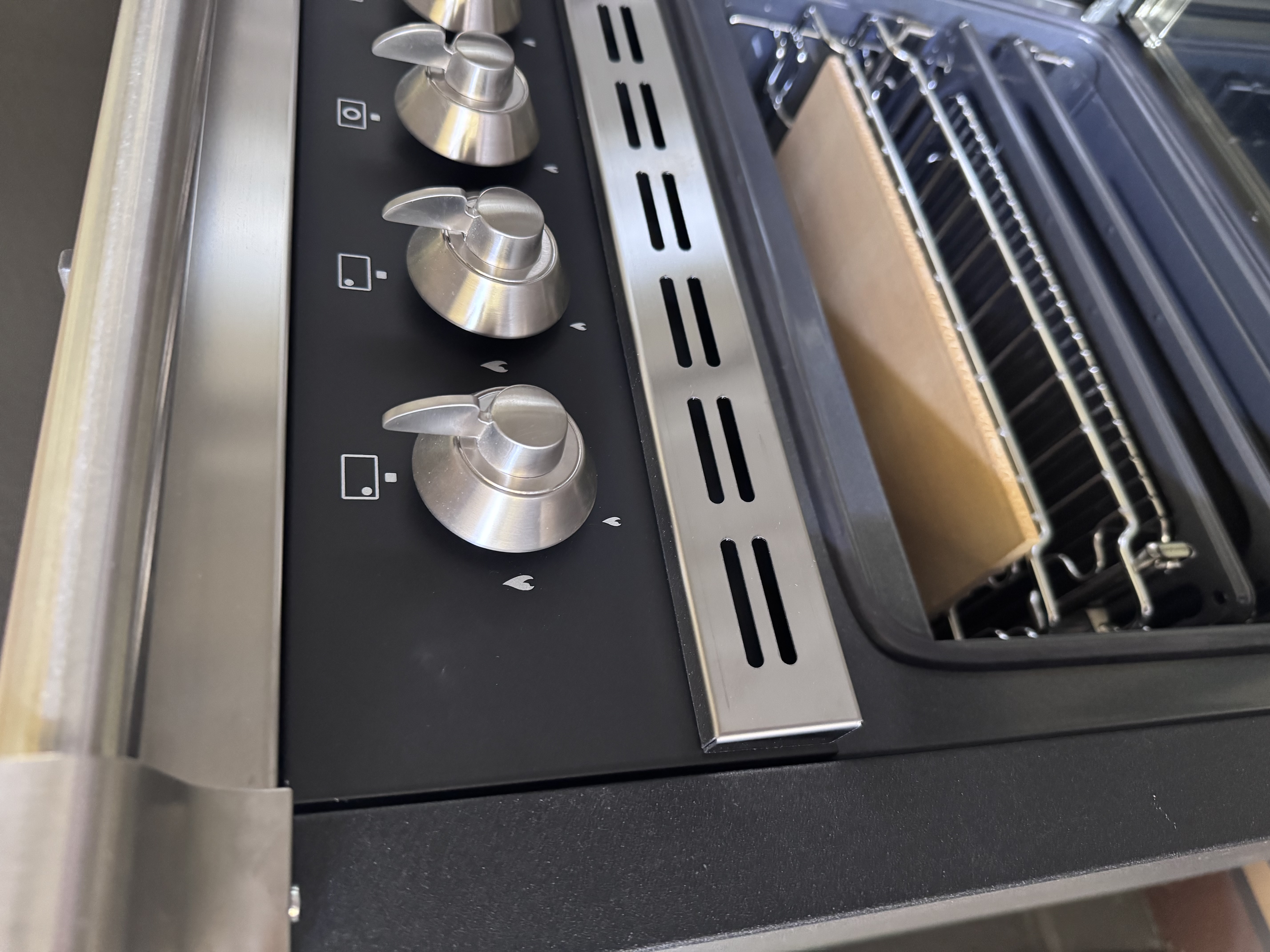 Nahaufnahme eines schwarzen Gasherds: vier Edelstahl-Drehknöpfe, darunter geöffneter Backofen mit Rost und Pizzastein