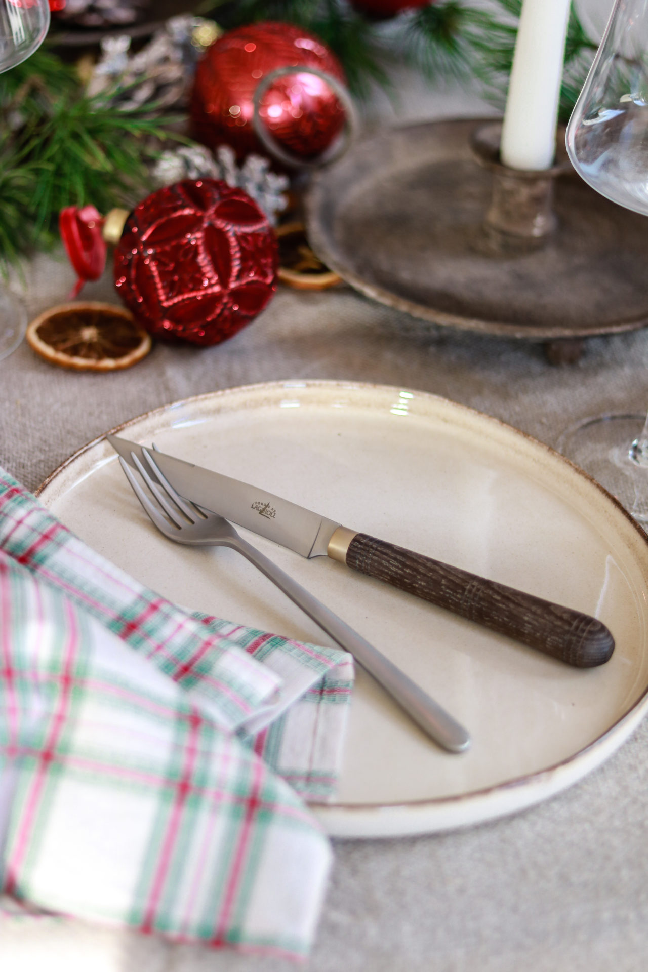 Festliche Tischdekoration mit Besteck, Kerze und Weihnachtskugel Ein gedeckter Weihnachtstisch mit gezacktem Messer und Gabel auf einem Teller, karierter Serviette, Kerze und roten Weihnachtskugeln als Dekoration