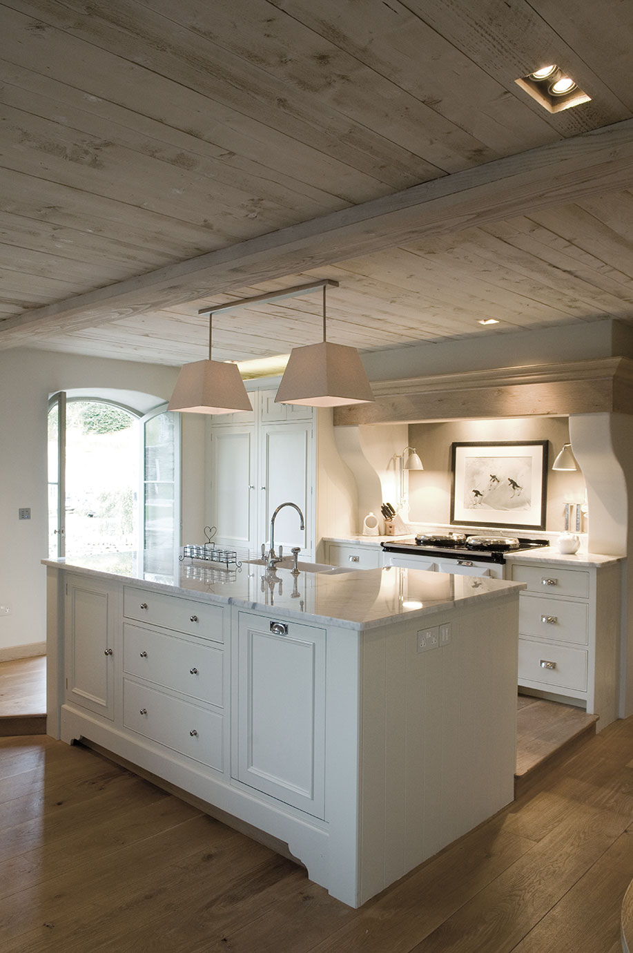 Moderne Landhausküche mit Kochinsel, hellen Fronten und Holzdecke Helle Landhausküche mit Küchenschrank, Kochinsel mit Marmorarbeitsplatte, zwei Hängeleuchten und Holzfußboden