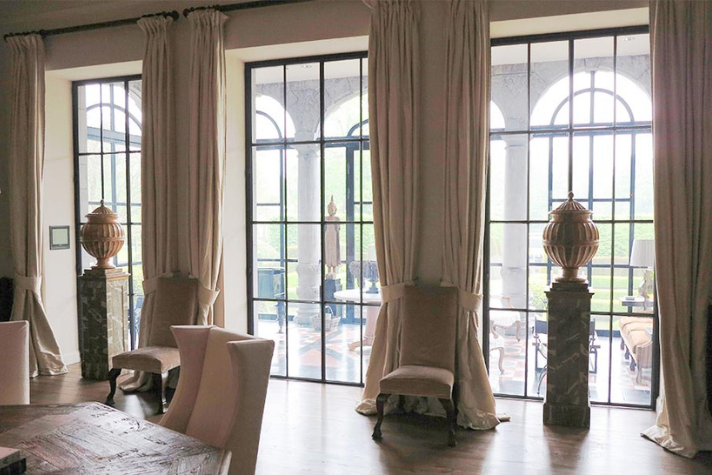 Großzügiger Wohnraum mit eleganter Einrichtung, bodentiefen Fenstern und klassischen Dekoelementen Heller Wohnraum mit hohen Vorhängen, Stühlen und großen, schwarzen Sprossenfenstern mit Blick auf eine Terrasse mit Säulen