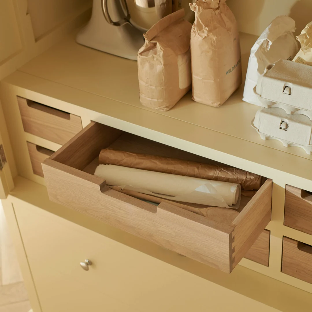 Offene Holzschublade in cremefarbenem Küchenschrank; darin Backpapierrollen und Löffel, oben Mehltüten und Eierkartons – zeigt übersichtliche Aufbewahrung im Küchenmöbel