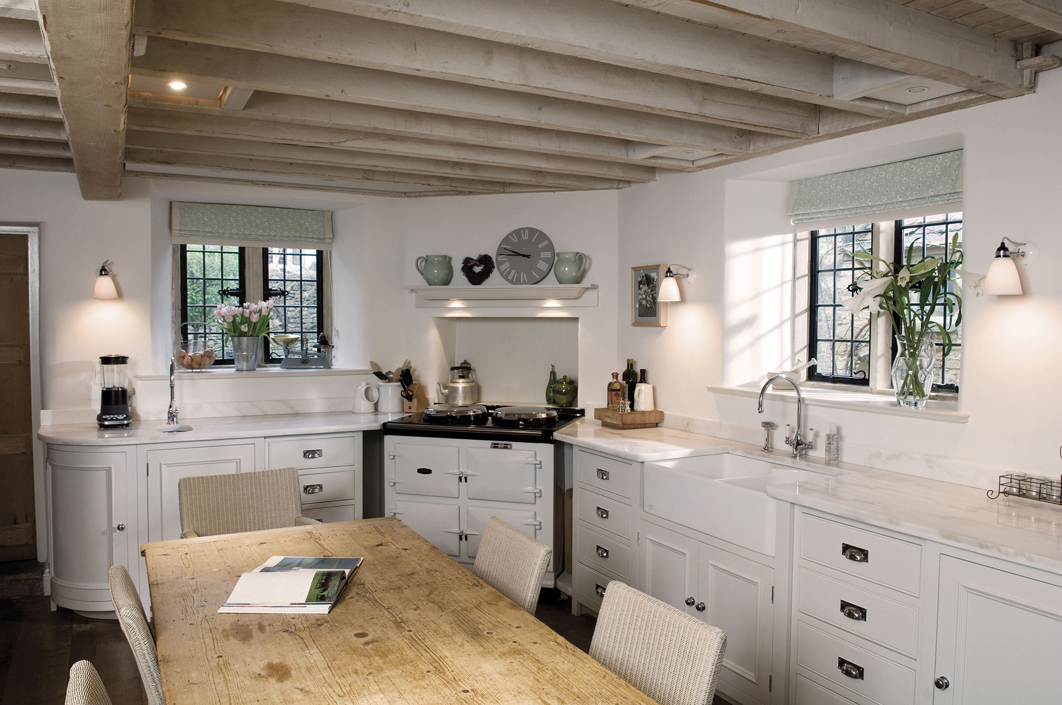 Helle Landhausküche mit Holztisch, weißen Schränken und großen Fenstern Gemütliche Landhausküche mit offenem Holzbalken-Decke, weißen Küchenmöbeln, Spüle, großem Holztisch und dekorativen Blumen auf der Fensterbank.