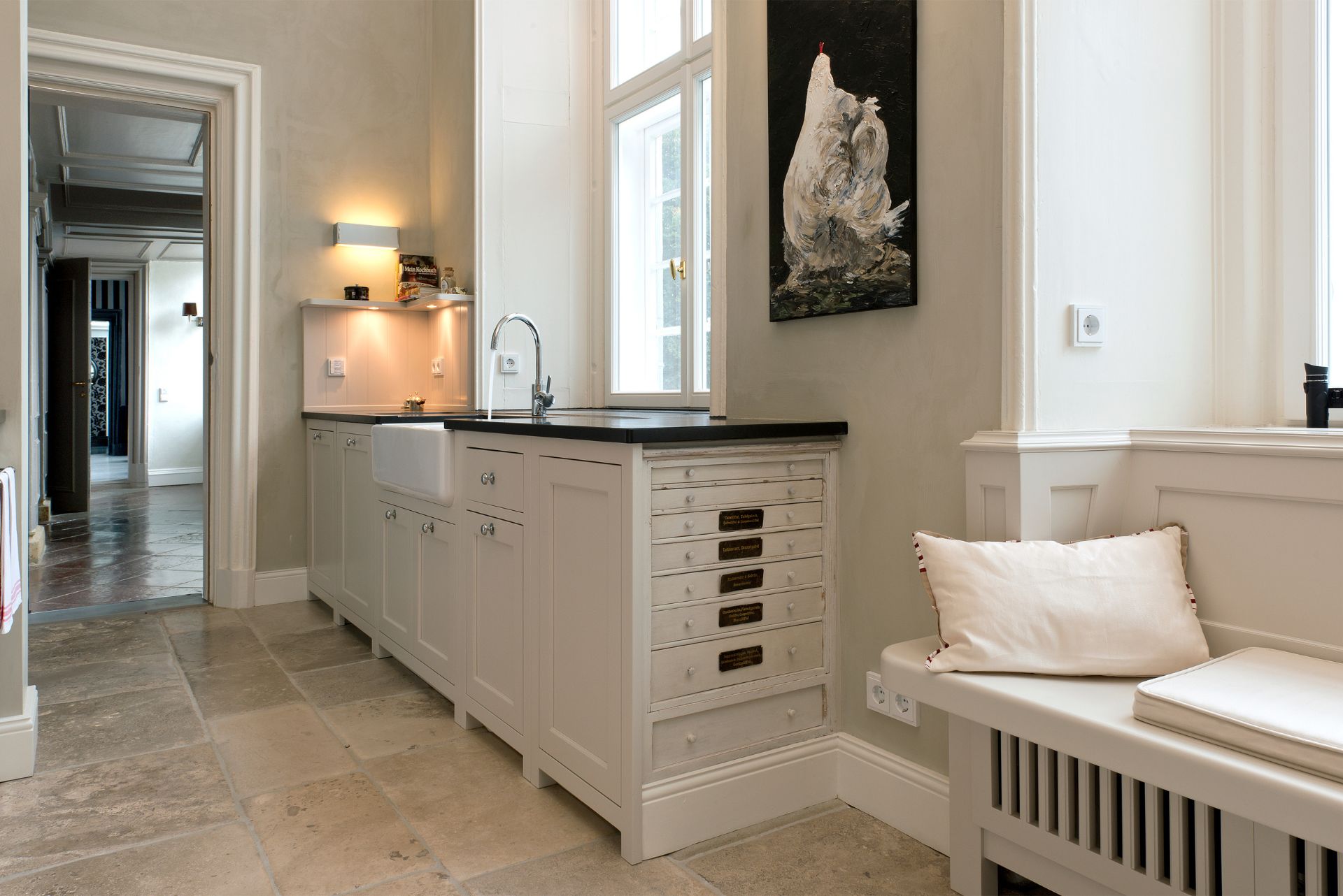 Helle Landhausküche mit Keramik-Spülbecken, weißen Schränken, schwarzer Arbeitsplatte, Natursteinboden und Sitzbank mit Kissen