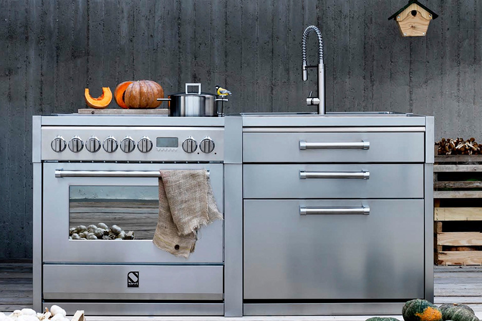 Moderne Outdoor-Küche aus Edelstahl mit Herd, Spüle und Arbeitsfläche, dekoriert mit Kürbis und Kochtopf Edelstahl-Outdoor-Küche mit Herd, Backofen, Spülbecken und Arbeitsfläche, darauf Kürbis, Topf und Küchentuch