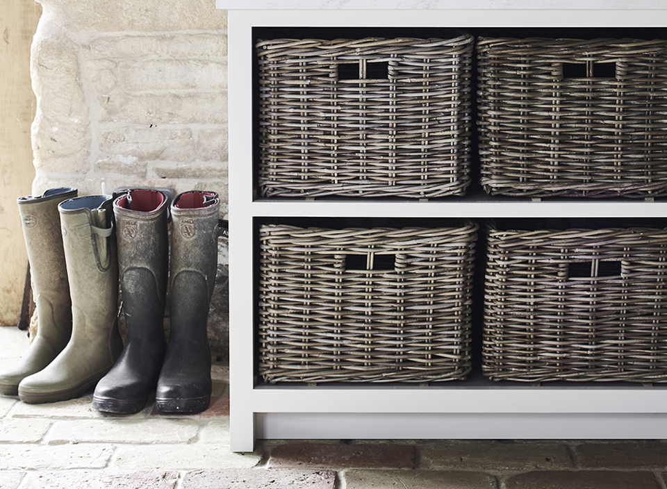 Garderobenschrank mit vier Weidenkörben und drei Paar Gummistiefeln auf Steinboden vor Natursteinwand Weißes Regal mit vier geflochtenen Weidenkörben, daneben drei Paar Gummistiefel auf einem Steinboden, rustikales Ambiente