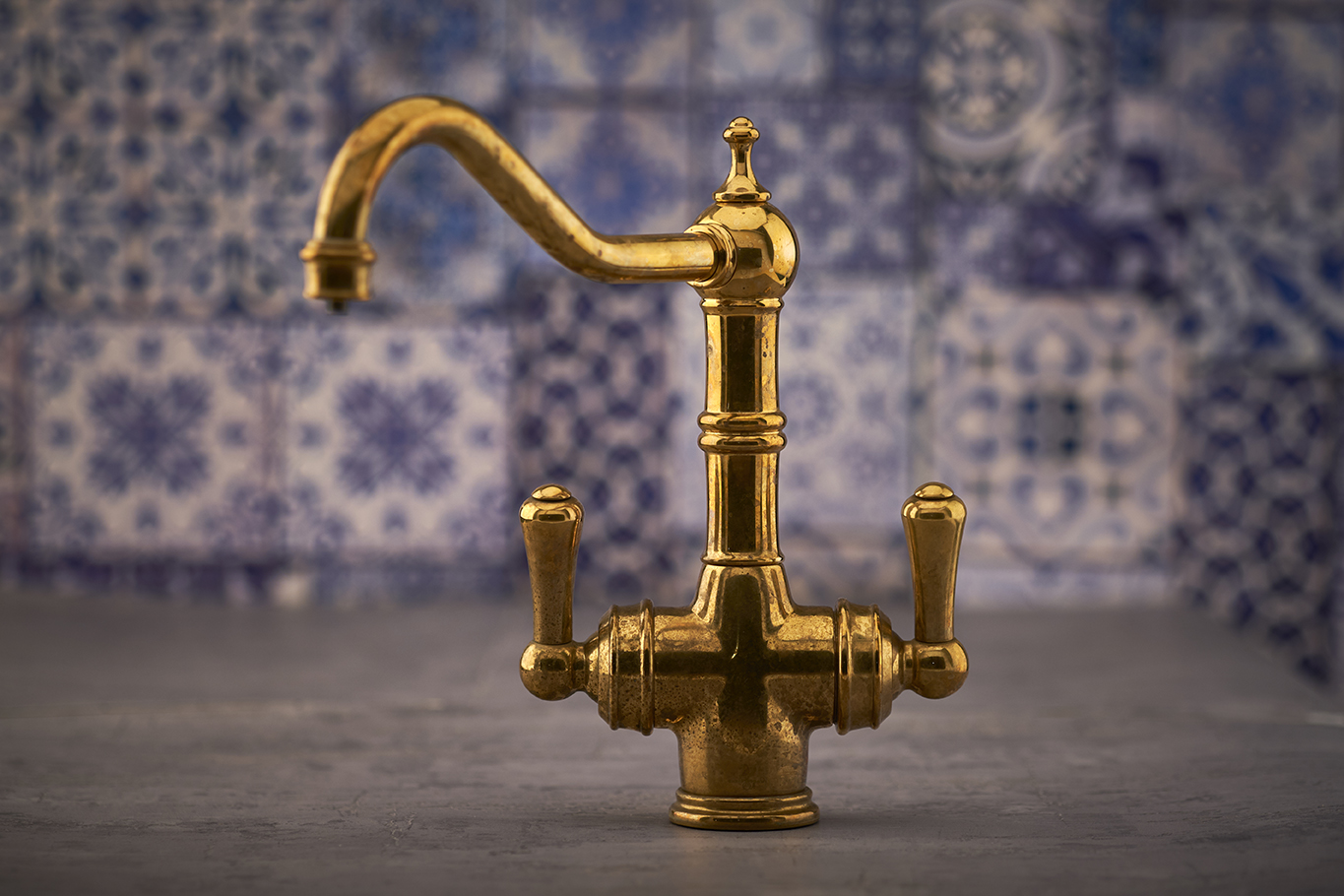 Antiker Wasserhahn aus Messing mit zwei Griffen und gebogenem Auslauf, ideal für nostalgische Küchen oder Bäder Goldfarbener Vintage-Messing-Wasserhahn mit zwei Griffen und gebogenem Auslauf vor gemusterter Kachelwand