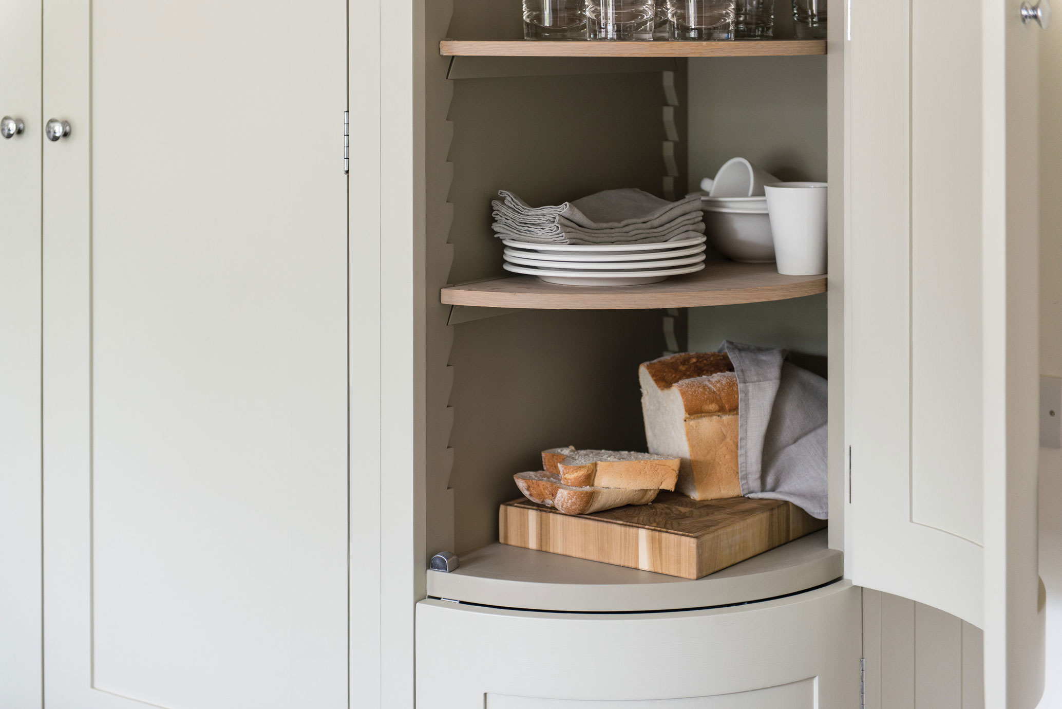 Offener Küchenschrank mit Brotschneidebrett, frischem Brot, Tellern, Gläsern und Tassen Ein geöffneter Küchenschrank mit gestapelten Tellern, Stoffservietten, Gläsern, Tassen sowie einem Schneidebrett mit frischem Brot und Brotscheiben