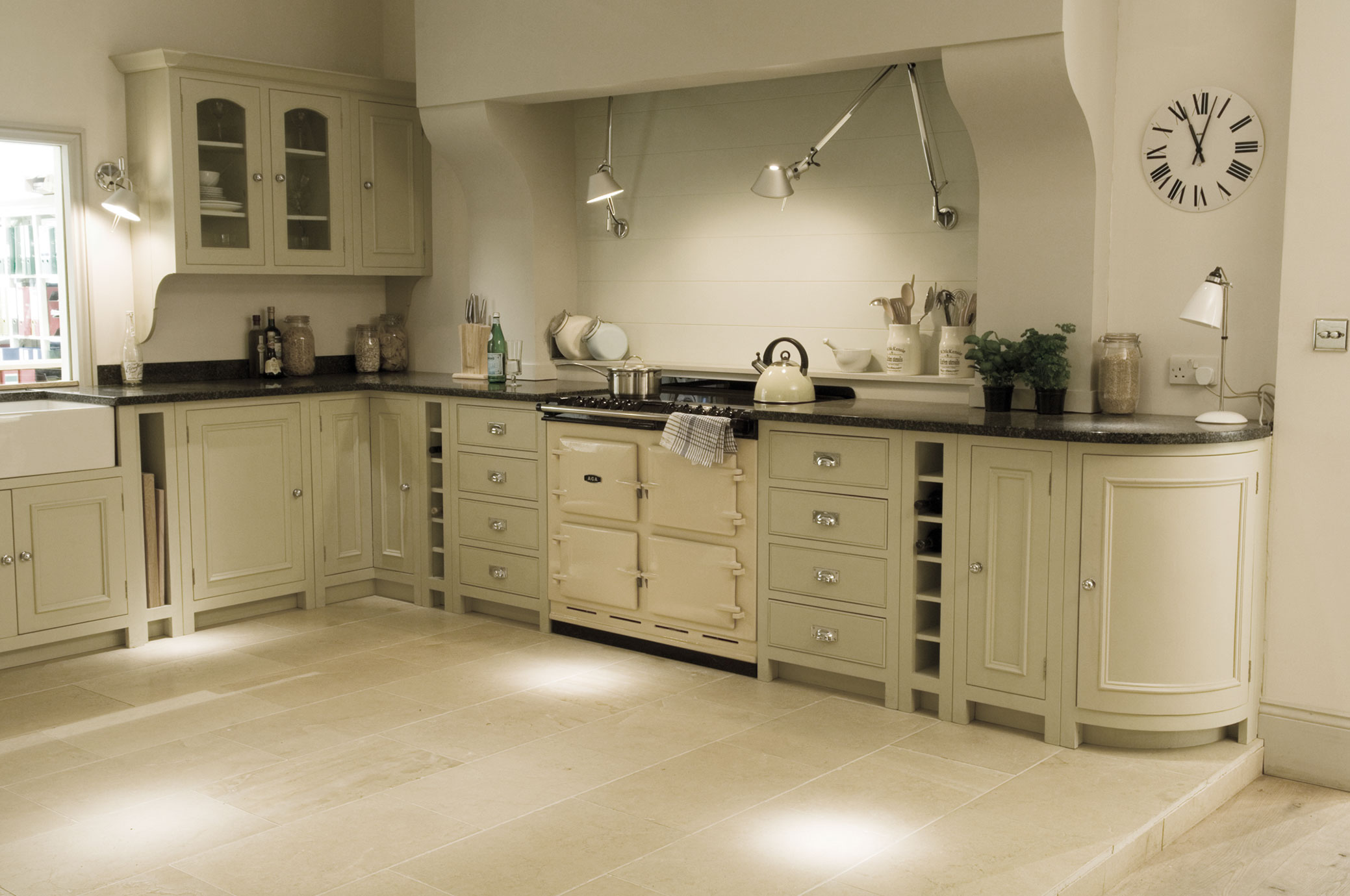 Moderne Landhausküche mit cremefarbenen Schränken, großem Herd und stilvollen Einbaulampen Helle Landhausküche mit cremefarbenen Küchenmöbeln, großem Herd, Wanduhren und offenem Stauraum auf beige gefliestem Boden