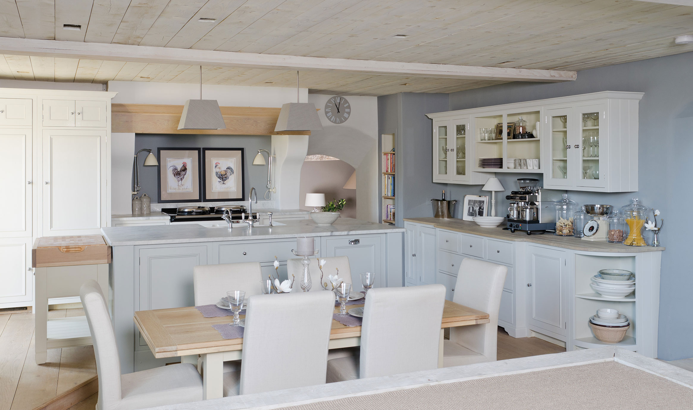 Moderne Landhausküche mit Essbereich, weißen Möbeln und stilvollen Accessoires Helle Landhausküche mit weißen Schränken, Essplatz für sechs Personen, Kücheninsel, Wandregalen und dekorativen Elementen