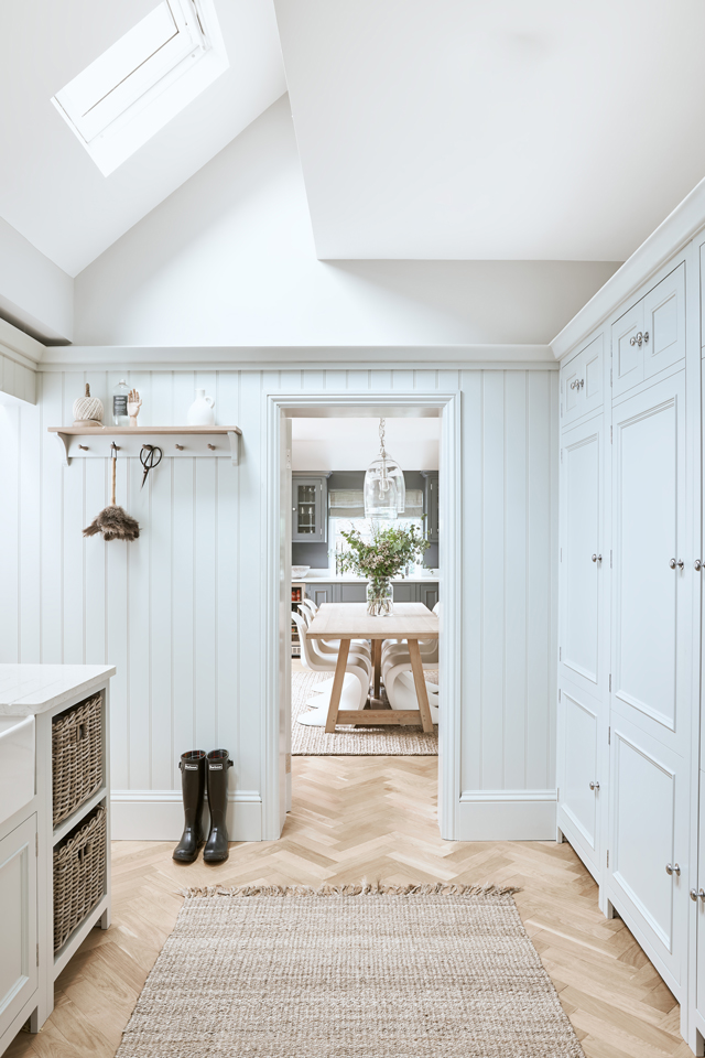 Helle, moderne Landhaus-Garderobe mit Blick in einen Essbereich Lichtdurchfluteter Flur im Landhausstil mit hellen Einbauschränken, gewebtem Teppich, Regalen, schwarzen Gummistiefeln und Blick auf einen gedeckten Esstisch mit Blumenvase.
