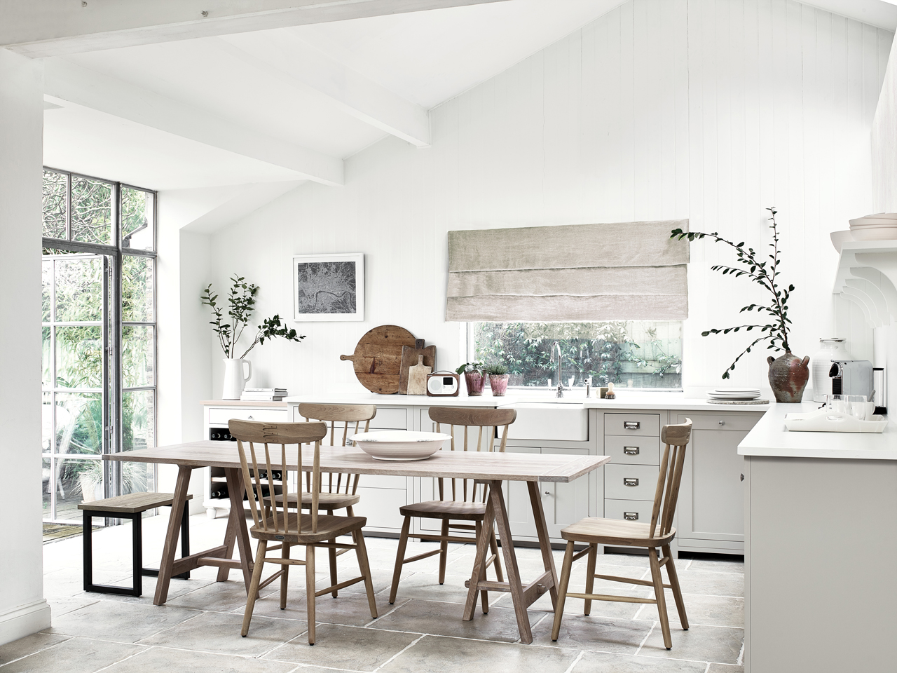 Helle, moderne Landhausküche mit Holztisch und Stühlen, großen Fenstern und minimalistischer Dekoration Lichtdurchflutete Küche im Landhausstil mit Holz-Esstisch, vier Holzstühlen, großen Fenstern, weißen Fronten und dekorativen Zweigen in Vasen