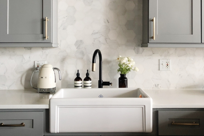 Moderne Küche mit grauen Schränken, weißer Spüle und hexagonaler Marmor-Rückwand Helle Küchenzeile mit grauen Schränken, weißer Landhausspüle, Wasserhahn in Schwarz, Wasserkocher, zwei Seifenspendern und Vase mit weißen Blumen