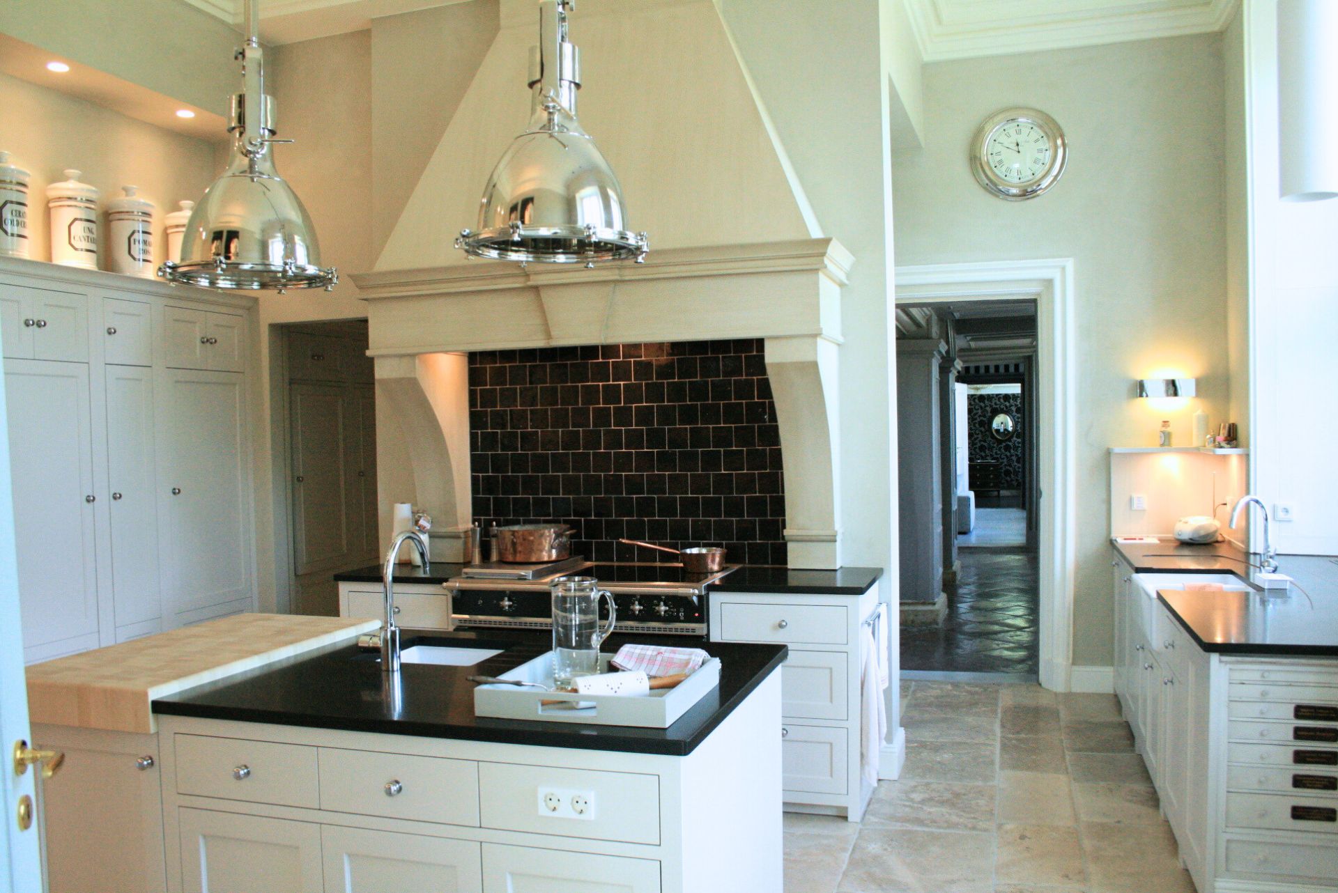 Helle Premium-Küche mit Kochinsel, schwarzer Stein-Arbeitsplatte, großem Herd vor schwarzer Fliesenrückwand, weißen Shaker-Fronten und zwei Chrom-Pendelleuchten – stilvolle Einrichtungsidee