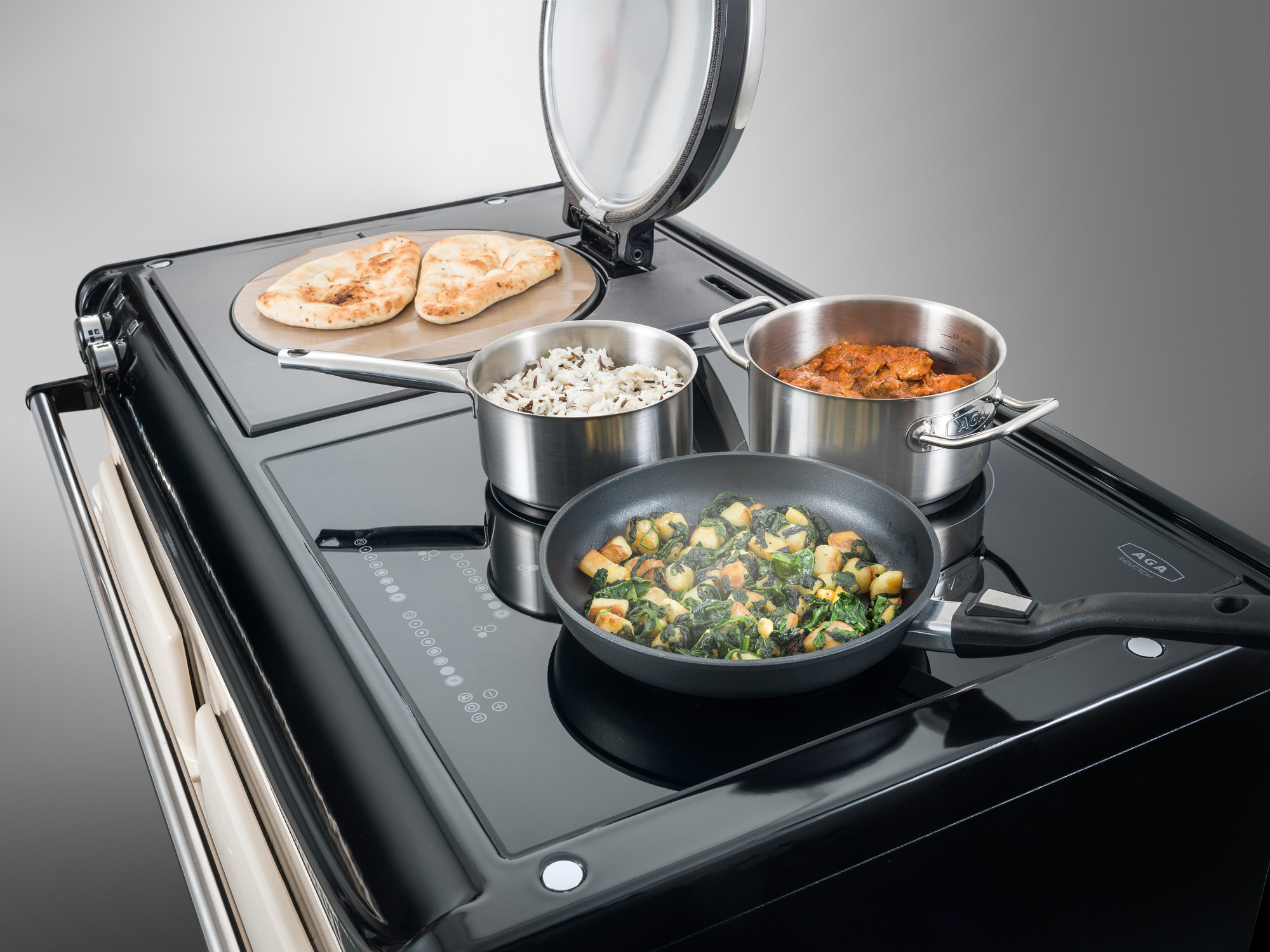 Moderner schwarzer Herd mit Induktionskochfeld, darauf stehen Edelstahl-Töpfe mit Reis, Curry, Gemüsepfanne und Fladenbrot Ein Induktionsherd mit drei Töpfen bzw. Pfannen: einer mit Reis, einer mit Curry, eine Pfanne mit Gemüse sowie Fladenbrot nebenan auf dem Herd