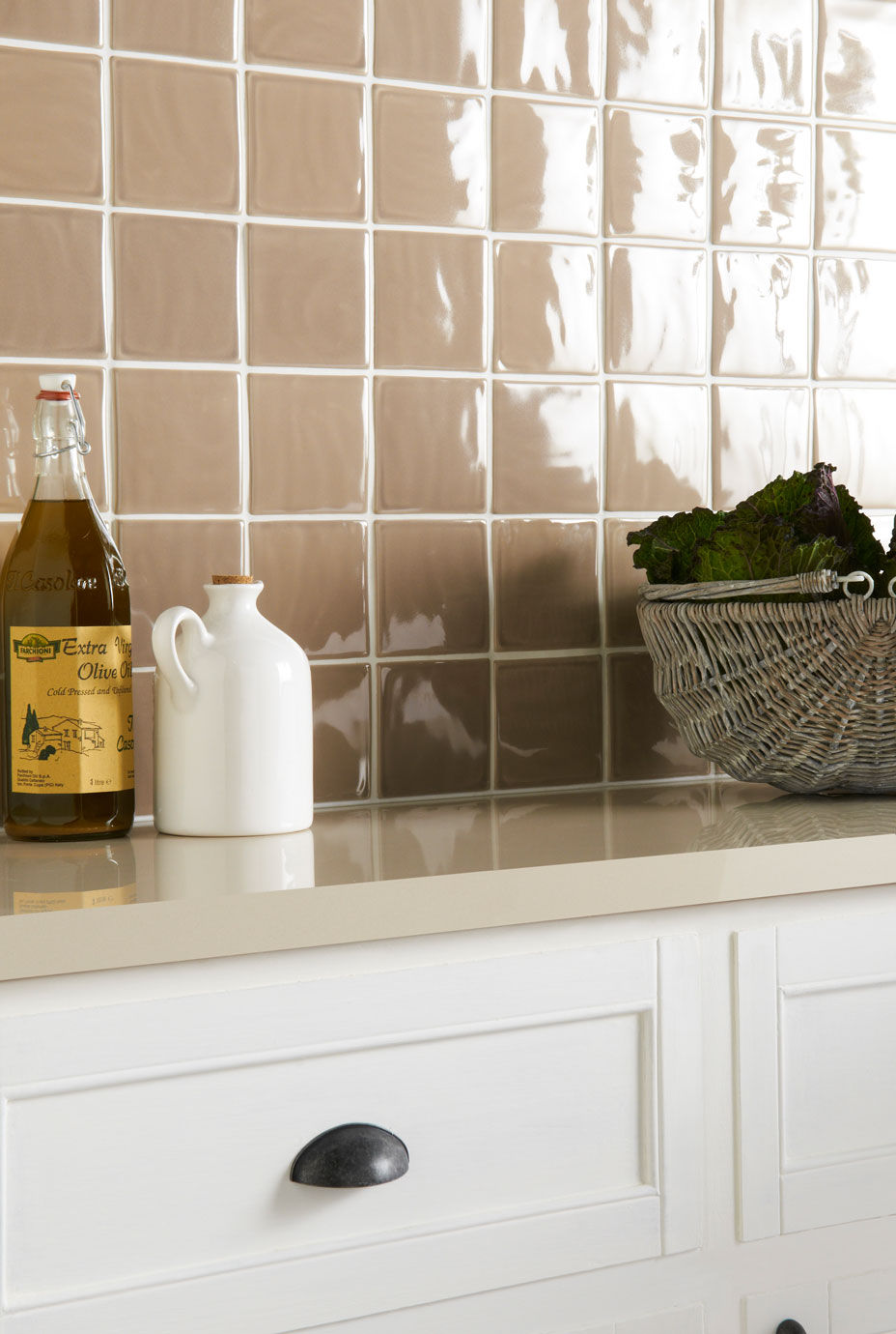 Küchenrückwand mit glänzenden, beigen Fliesen und moderner Arbeitsplatte Detailaufnahme einer Küche mit glänzenden, beigen Wandfliesen, Flasche Olivenöl, weißem Keramikkrug und Korb mit Gemüse auf hell beiger Arbeitsplatte
