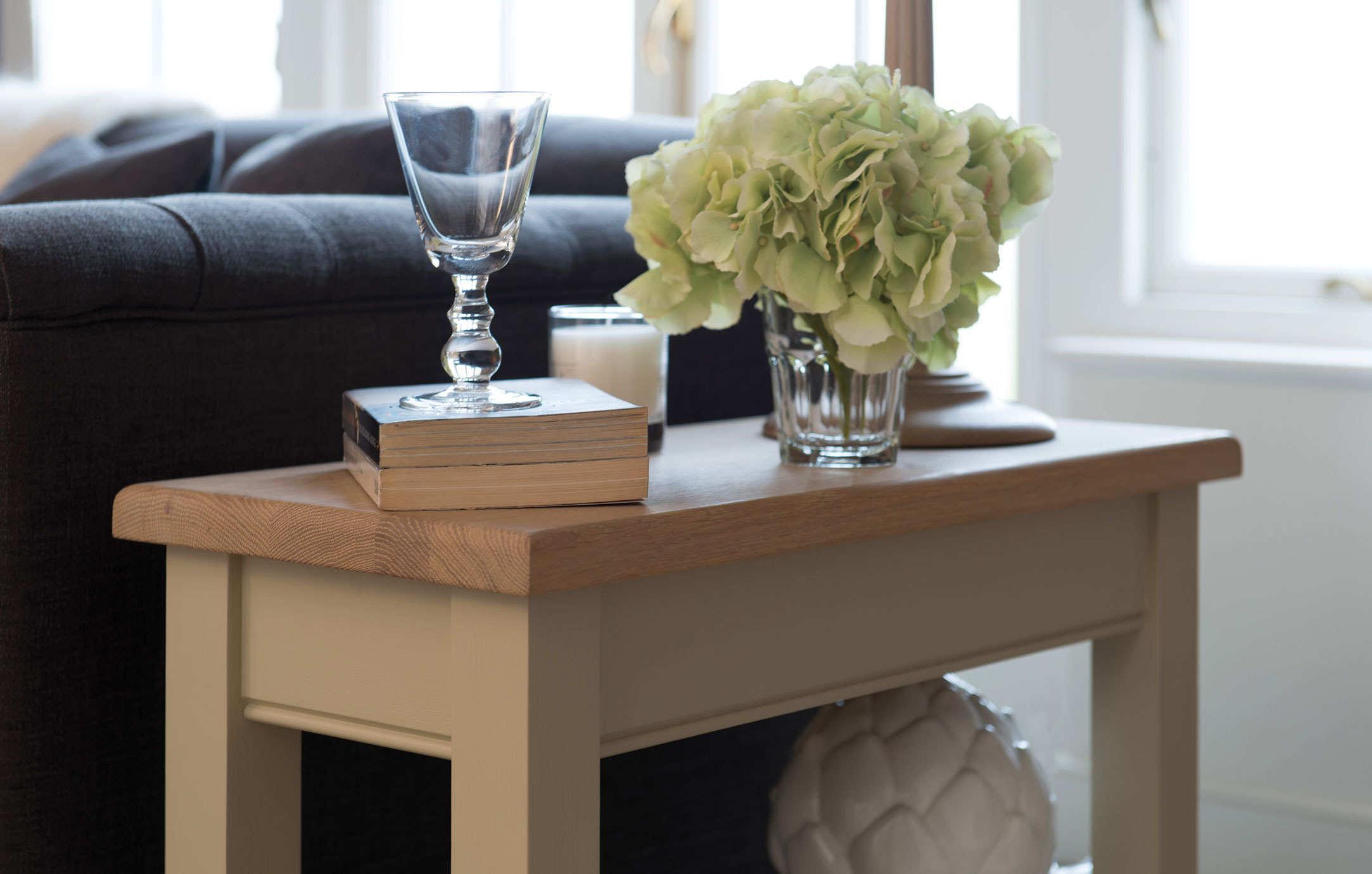 Beistelltisch aus Holz mit Dekoration im Wohnzimmer Beistelltisch mit Holzplatte, darauf ein Weinglas, Bücher, eine Kerze und ein Blumenstrauß, daneben ein graues Sofa im Wohnzimmer