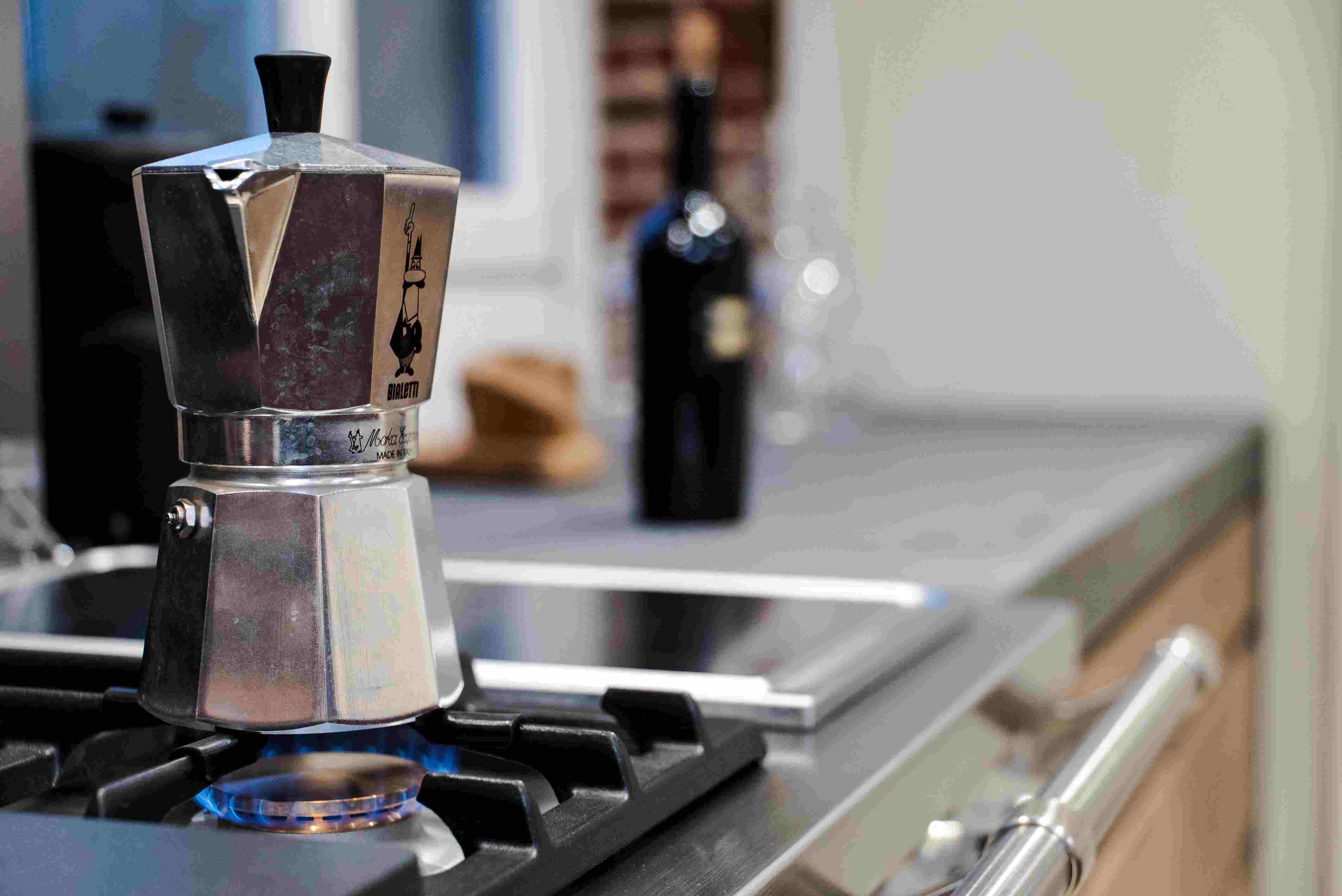 Ein Espressokocher auf einer Gasflamme in einer modernen Küche, im Hintergrund eine Flasche und ein Hut