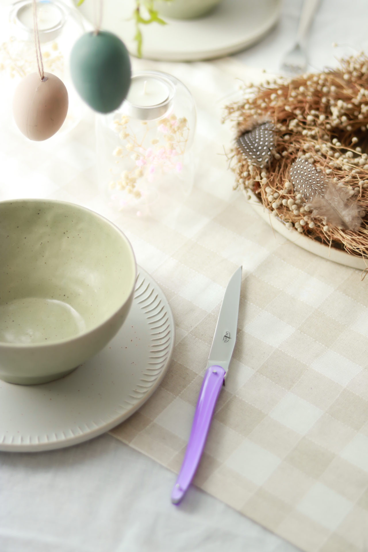 Festlich gedeckter Ostertisch mit Keramikschale, Nest und Messer mit lila Griff Detail eines österlich dekorierten Tisches mit pastellfarbener Keramikschale, Messer mit lila Griff, Deko-Eiern, Kerze und geflochtenem Nest mit Federn