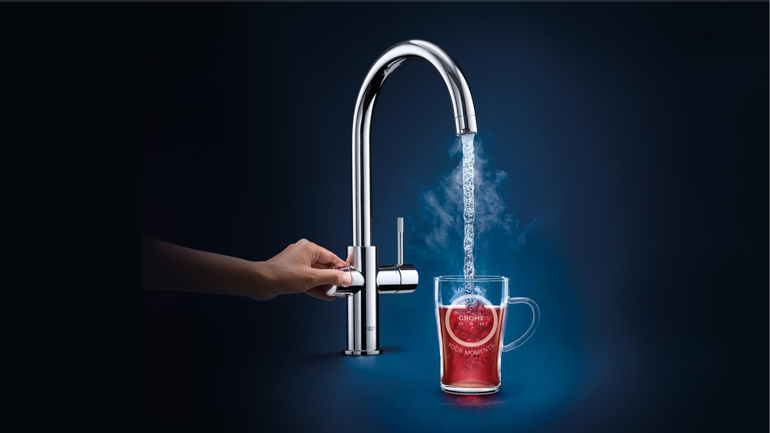 Moderne Küchenarmatur mit Heißwasserfunktion – Grohe Wasserhahn für sofort kochend heißes Wasser Hand bedient einen Grohe Wasserhahn, der kochend heißes Wasser in ein Teeglas gießt, aus dem Dampf aufsteigt