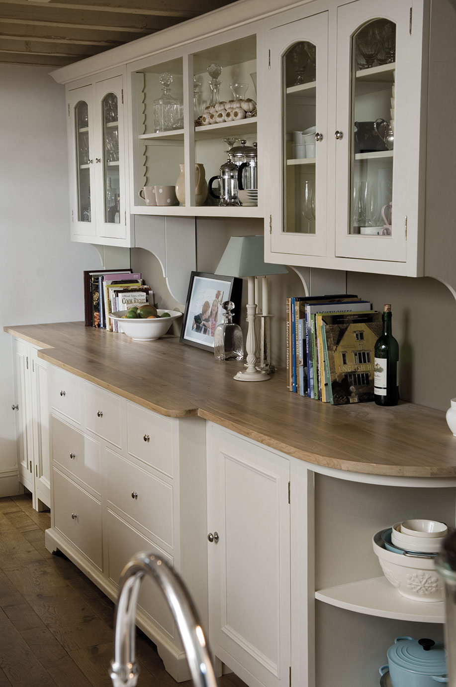 Eleganter Küchen-Unterschrank mit Holzarbeitsplatte und Vitrinenaufsatz in weißem Landhausstil Weißer Küchenunterschrank mit Holzarbeitsplatte, Glastürenschränken, Kochbüchern, Geschirr, Schalen und Flaschen in einer stilvollen Landhausküche