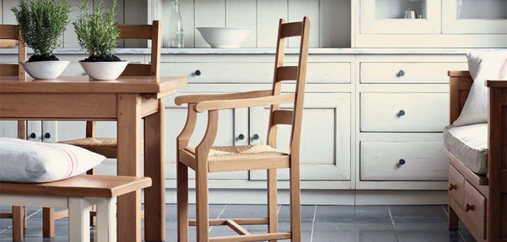Esszimmer aus Holz mit Tisch, Stühlen und Bank vor weißer Landhausküche Freundliches Esszimmer mit Holztisch, Stühlen, Bank und weißer Landhausküche im Hintergrund. Pflanzen als Dekoration auf dem Tisch.