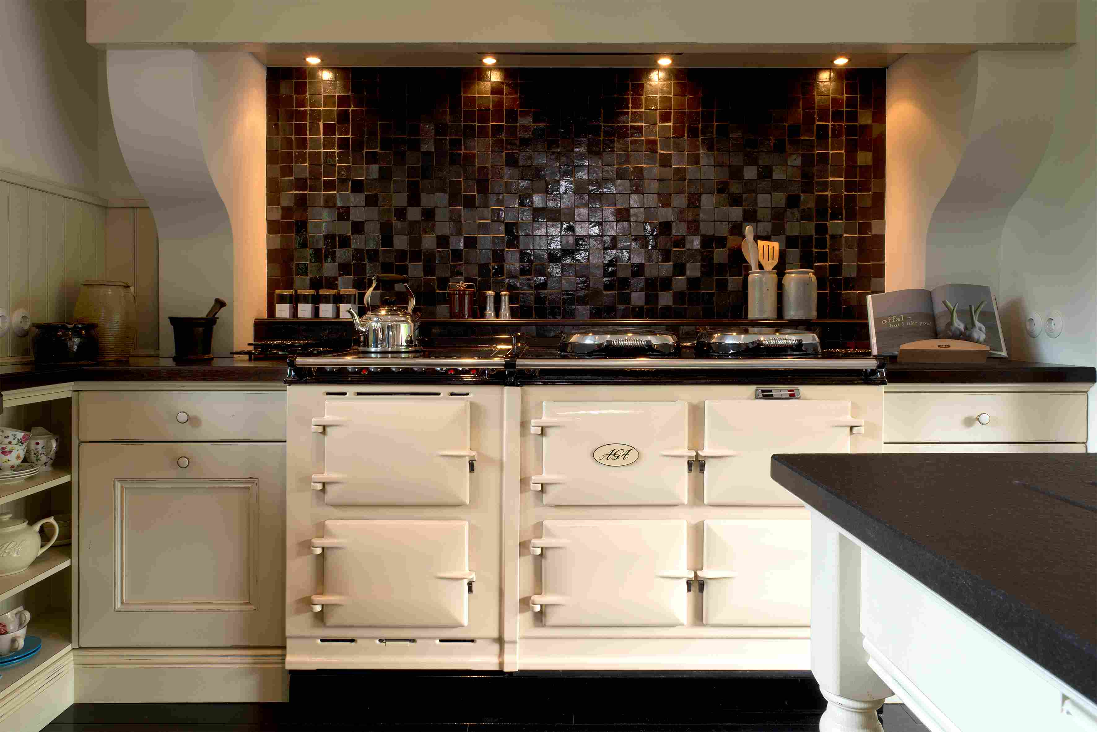 Klassischer cremefarbener AGA-Herd in moderner Küche mit schwarzer Mosaikfliesen-Rückwand Eleganter, cremefarbener AGA-Küchenherd vor einer schwarzen Mosaikfliesen-Rückwand, beleuchtet durch Einbauleuchten. Auf der Arbeitsfläche stehen eine Teekanne, Küchenutensilien und Gewürze.