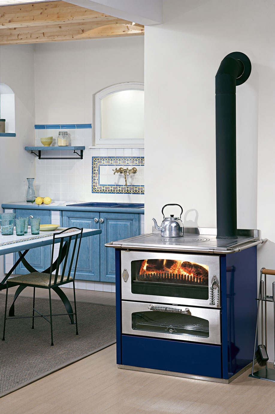 Moderner blauer Holzofen mit Edelstahl-Oberfläche in einer gemütlichen Landhausküche mit blauen Schränken Gemütliche Küche im Landhausstil mit blauem Holzofen, sichtbarem Feuer, Teekessel und Küchenmöbeln in Blau