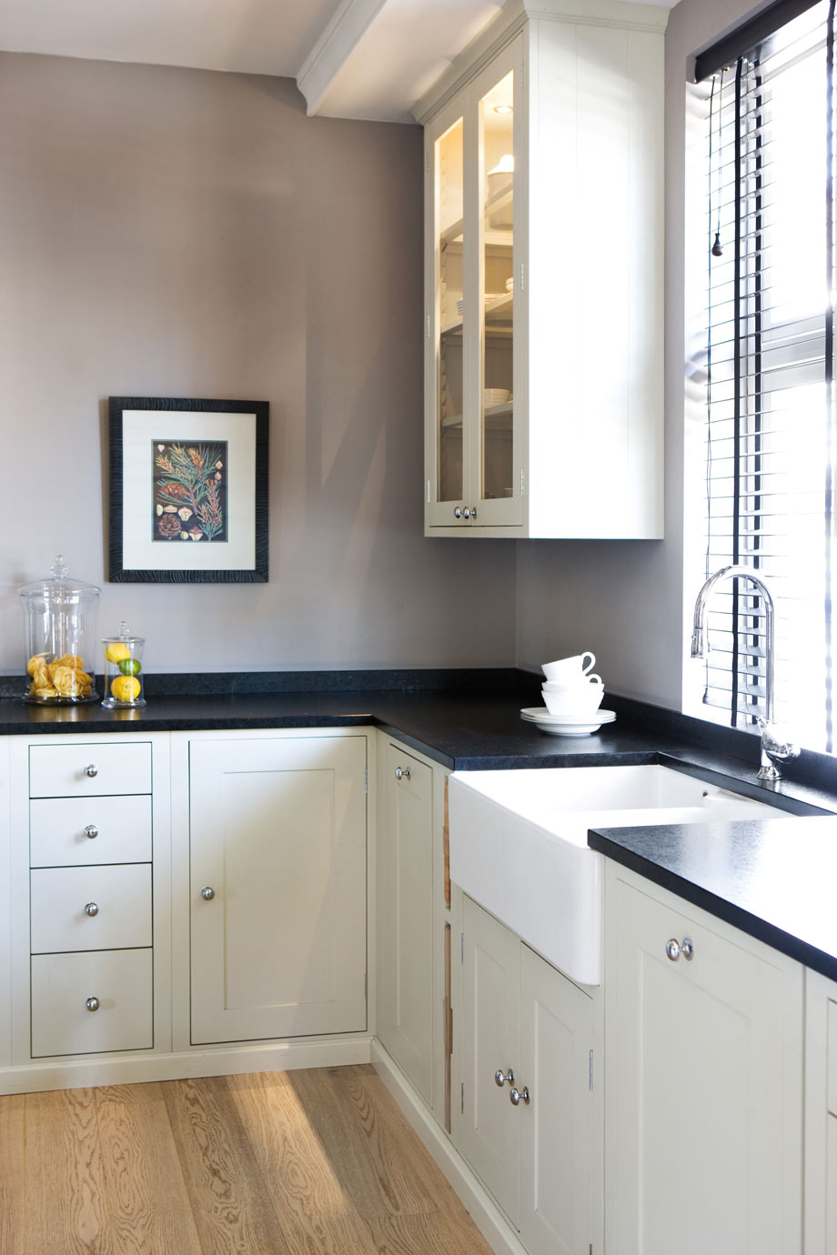 Moderne helle Küche mit schwarzen Arbeitsplatten und Landhausstil-Spüle Helle Einbauküche mit schwarzen Arbeitsplatten, Landhausstil-Spüle, Glashängeschrank, Holzfußboden und großen Fenstern mit Lamellenjalousien