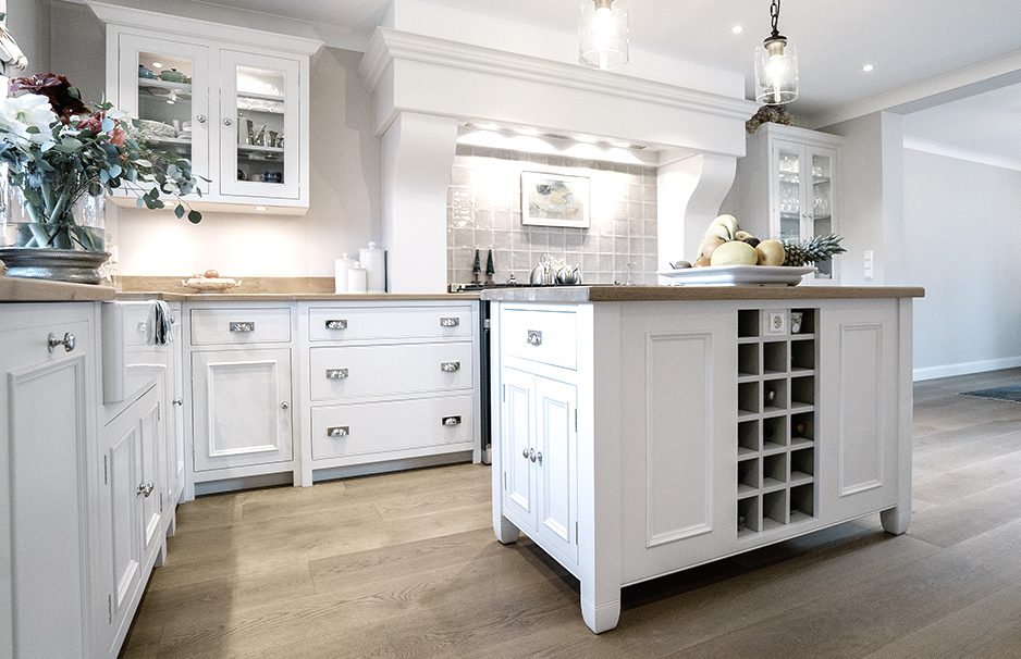 Moderne Landhausküche mit Kochinsel, weißen Schränken und Holzboden Helle, moderne Landhausküche mit weißer Kochinsel, integriertem Weinregal, Obstschale und großzügigem Stauraum auf hellem Holzboden