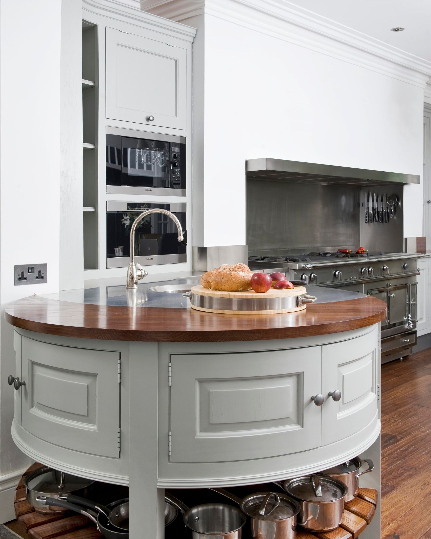 Moderne Küche mit runder Kücheninsel, Holzarbeitsplatte und Edelstahlherd Helle Designerküche mit runder Kücheninsel aus Holz und Edelstahl, Brotlaib und Äpfeln auf einem Schneidebrett sowie mehreren Töpfen im unteren Regalbereich