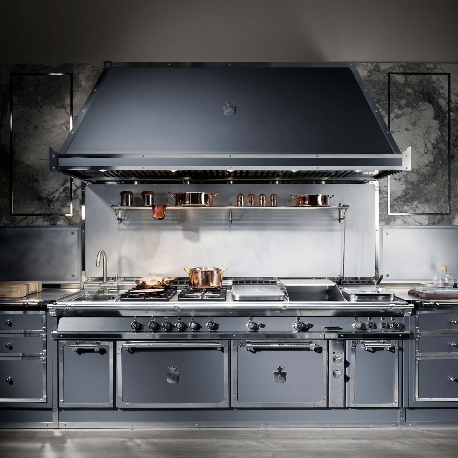 Hochwertige Edelstahl-Kochstation mit großer Dunstabzugshaube, mehreren Gasbrennern, Plancha/Grillplatte und Backöfen; Kupfer-Töpfe auf dem Herd – Produktfoto einer Premium-Profiküche