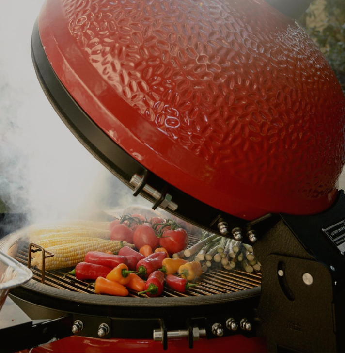 Keramikgrill mit buntem Grillgemüse wie Mais, Tomaten, Paprika und Spargel im Outdoor-Bereich Großer roter Keramikgrill mit geöffnetem Deckel, darin frisches Gemüse wie Maiskolben, Tomaten, Paprika und Spargel auf dem Grillrost, umgeben von Rauch