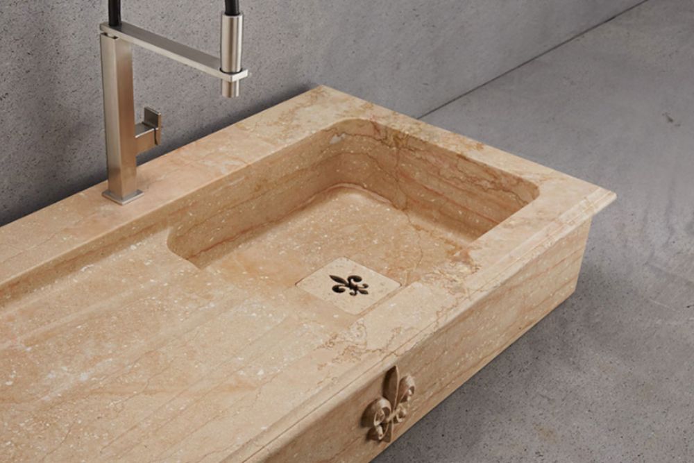 Beige Naturstein-Küchenspüle aus Marmor mit integrierter Abtropffläche, eckigem Becken und Edelstahlarmatur; Ablaufabdeckung und Front mit Lilien-Relief – massiver Spülstein für Landhausküchen