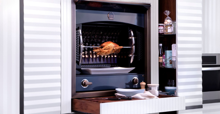 Moderner Grillofen mit einem Brathähnchen am Spieß in einer stilvollen Küche Ein Brathähnchen wird auf einem Drehspieß in einem modernen Grillofen in einer eleganten Küche zubereitet, daneben stehen Gewürze und Geschirr