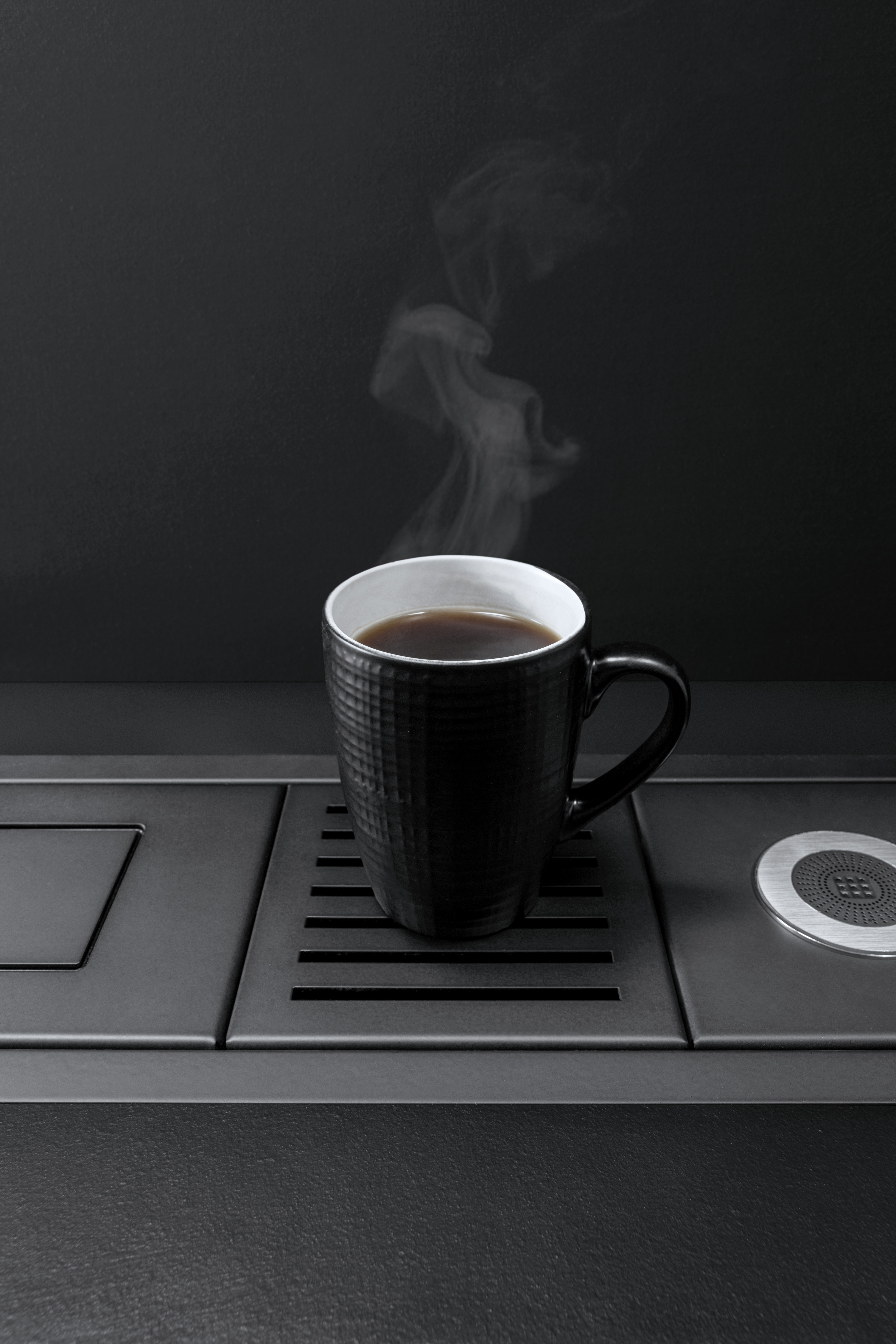 Schwarze Kaffeetasse mit heißem Kaffee auf moderner Küchenoberfläche Dampfende schwarze Kaffeetasse steht auf einer eleganten, dunklen Küchenoberfläche neben einem Abflussgitter
