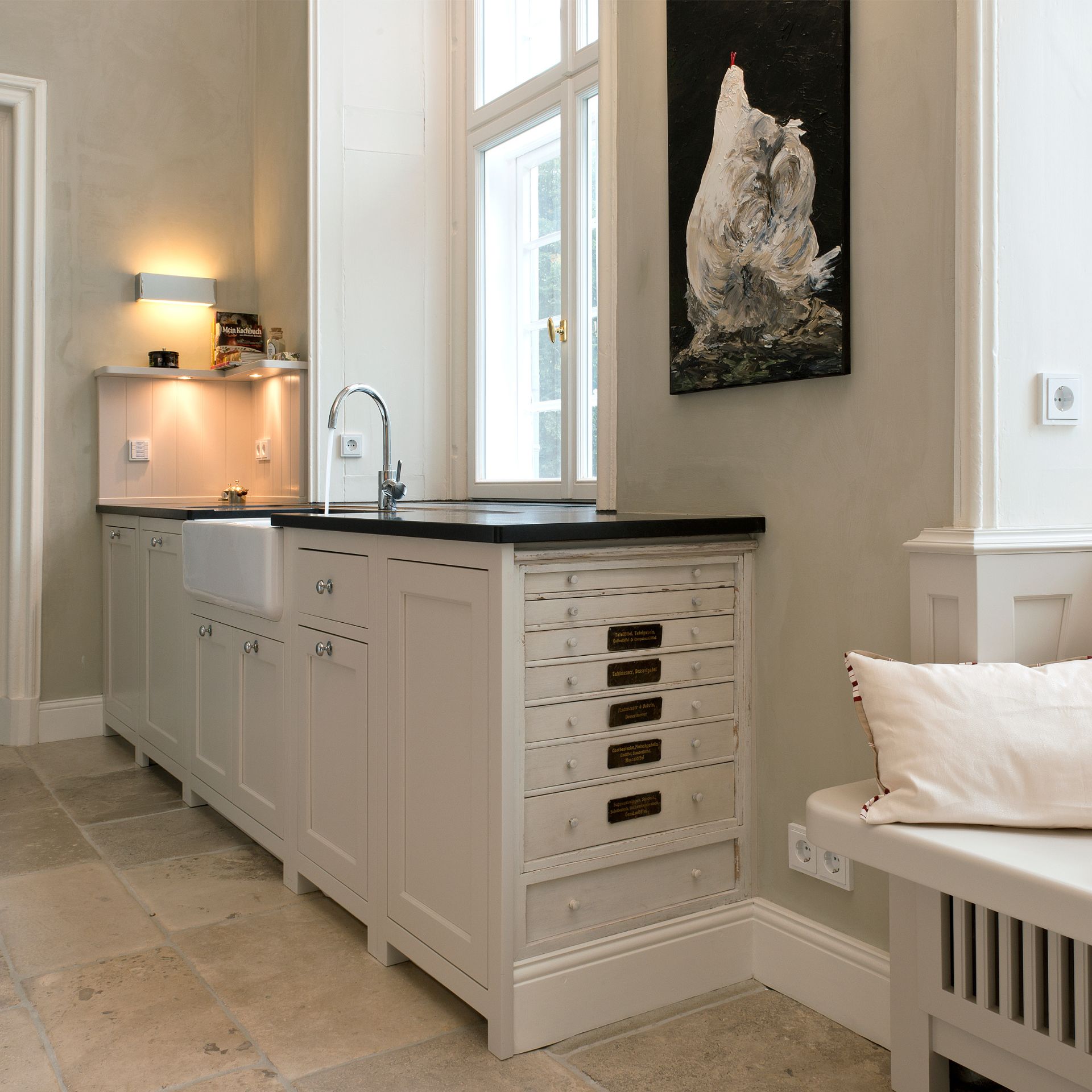 Helle Landhausküche: cremefarbene Shaker-Schränke mit Keramik-Spülbecken, schwarze Arbeitsplatte, Apothekerschubladen, Steinboden und Fenster – ideal zur Präsentation hochwertiger Küchenmöbel