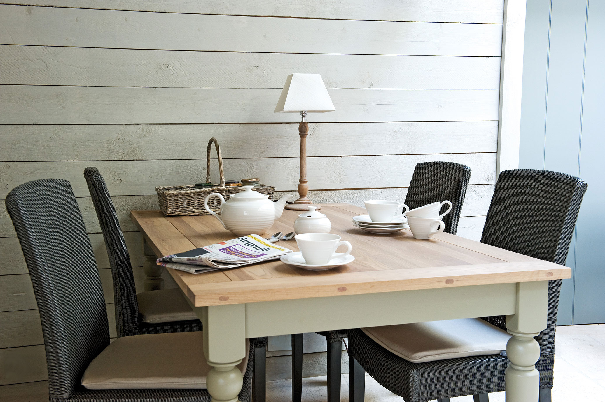 Esszimmer im Landhausstil mit Holztisch, vier Korbstühlen, Teeservice und Tischlampe Gemütliches Esszimmer mit Holztisch, vier Korbstühlen, einem weißen Teeservice, Zeitschrift, Tischlampe und Weidenkorb vor einer hellen Holzvertäfelung