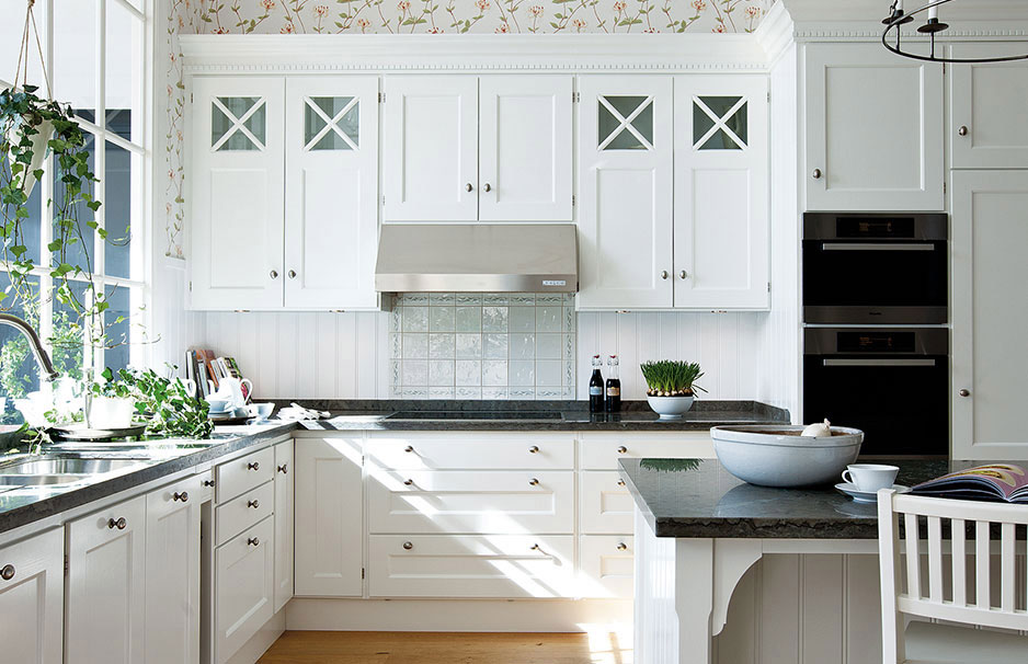 Moderne Landhausküche in Weiß mit eleganten Einbauschränken und dunkler Arbeitsplatte Helle, gemütliche Landhausküche mit weißen Schränken, großen Fenstern, Pflanzen und moderner Ausstattung