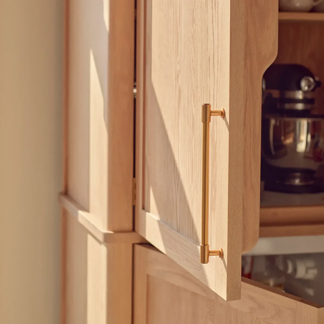 Detailansicht eines hellen Holz-Küchenschranks mit geöffneter Tür und langer Griffstange aus gebürstetem Messing; hochwertig verarbeitetes, modernes Schrankgriff-Design.