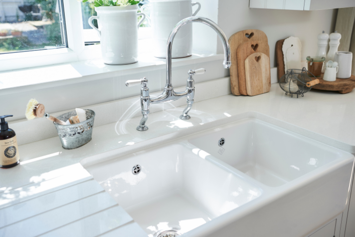 Moderne Doppelspüle in heller Landhausküche mit eleganter Armatur und dekorativen Küchenaccessoires Helle Doppelspüle mit glänzender Armatur in einer stilvollen Küche, umgeben von Küchenutensilien und frischen Pflanzen auf der Fensterbank