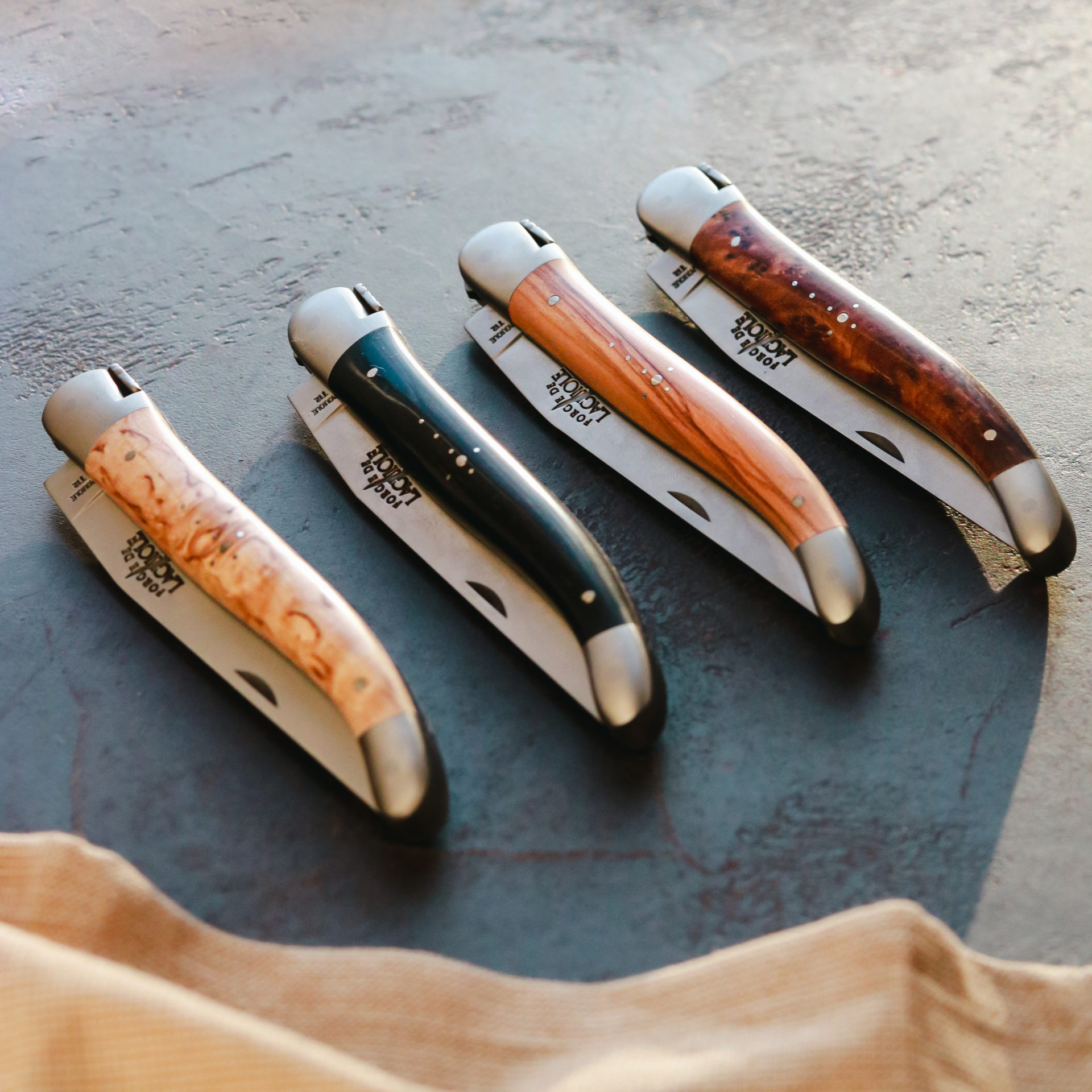 Elegante Taschenmesser mit Holzgriff – Set aus vier verschiedenen Modellen Vier stilvolle Taschenmesser mit unterschiedlichen Holzgriffen in einer Reihe auf dunkler Oberfläche.