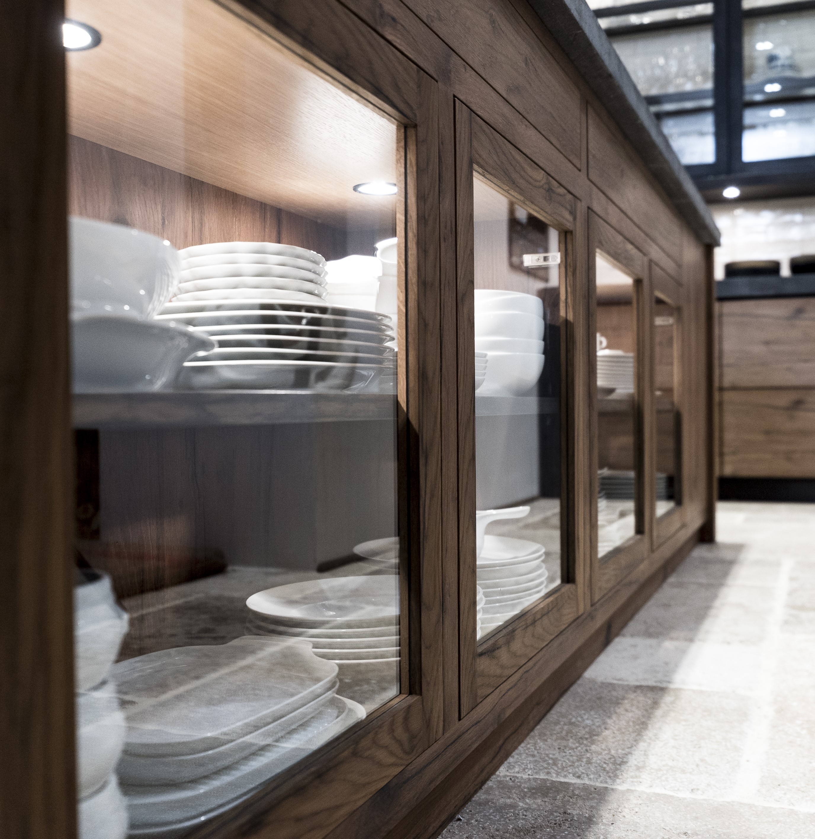 Holzküchenschrank mit Glasoberflächen und weißem Geschirr im Inneren, auf einem Steinboden stehend