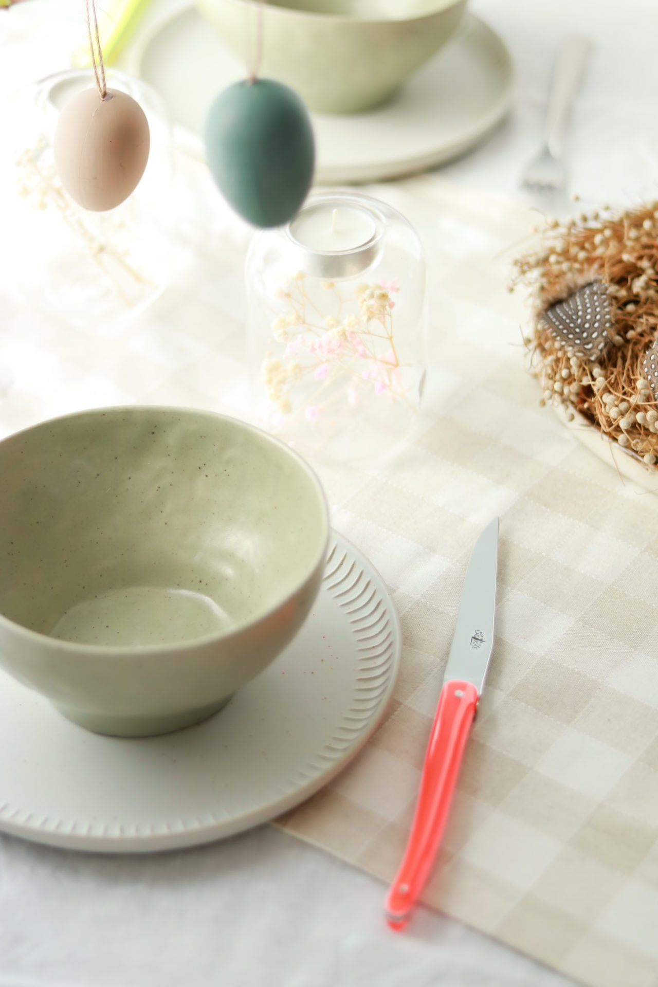 Frühlingshafte Tischdekoration mit Keramikschale, Ostereiern und Besteck auf kariertem Tischtuch Stilvoll gedeckter Tisch mit heller Keramikschale auf Teller, Messer mit pinkem Griff, dekorativen Ostereiern und Trockenblumen auf einem karierten Tischtuch